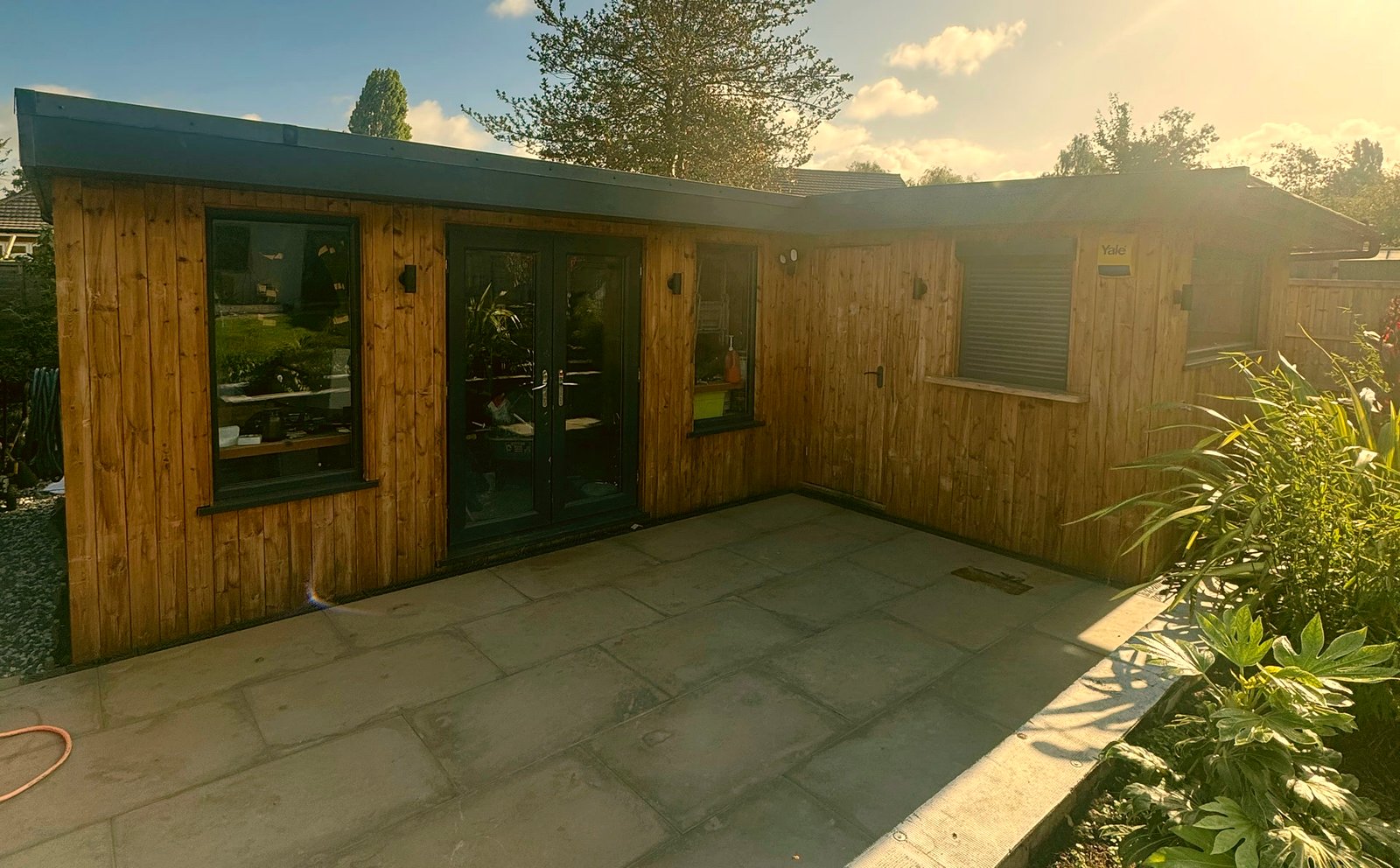 An L-shaped timber garden room with a paved patio and a raised bed, in sunshine